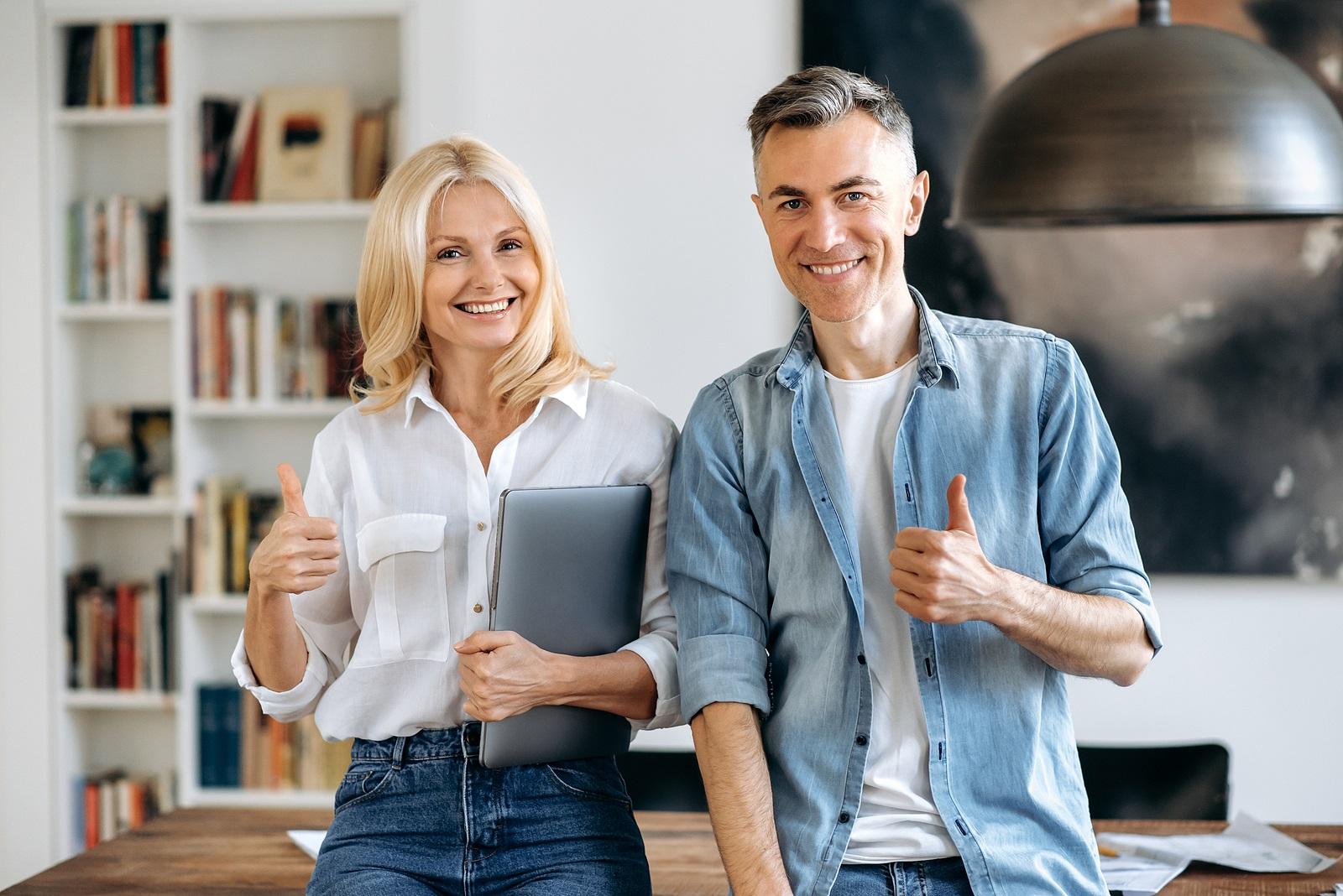 Portrait of two successful business colleagues. An elegant business lady and a confident business man, stand near a work desk in a modern office, look at the camera, show thumbs up gesture, smile